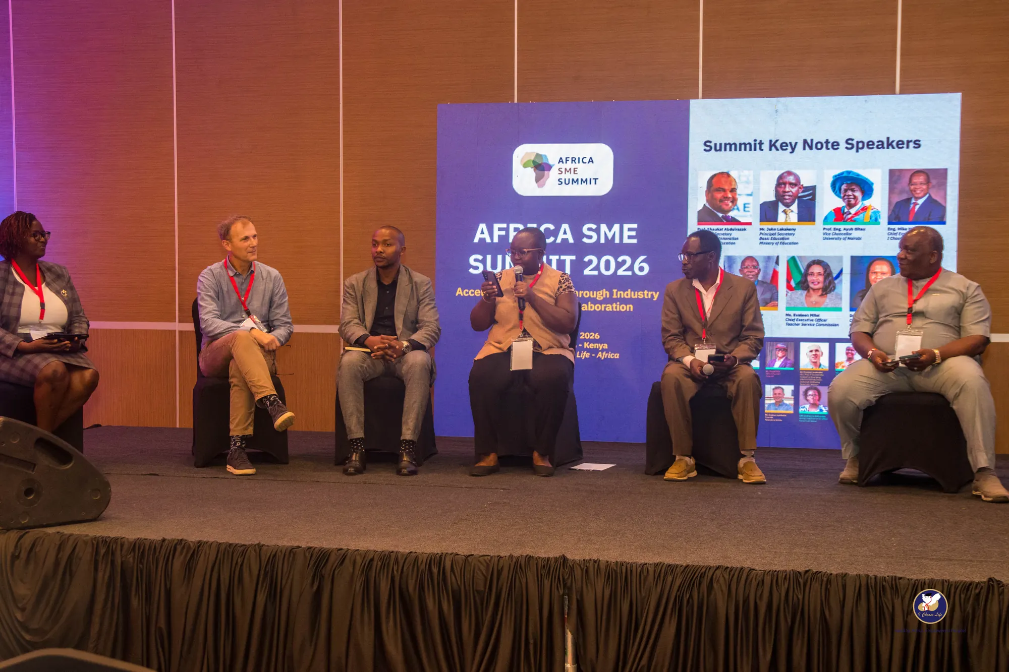 Panellists seated on stage in front of the screen listing the summit's keynote speakers