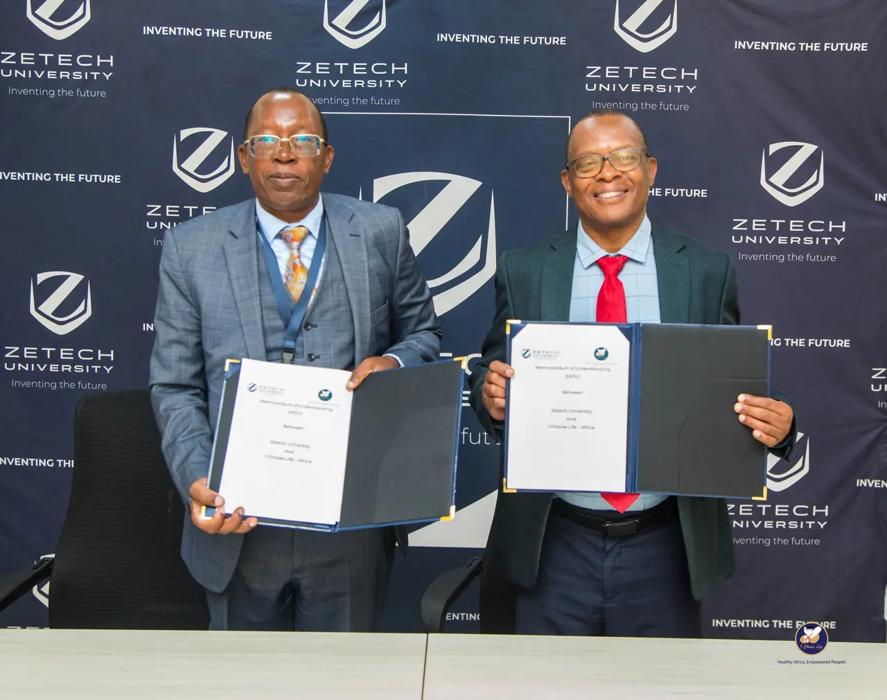 Eng. Mike Mutungi and Prof. Njenga Munene each hold open folders containing the signed MoU in front of the Zetech University event backdrop.